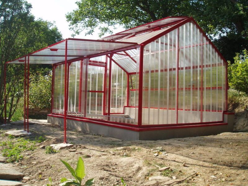 Gewächshaus mit rotem Rahmen und überdachter Veranda in einer Gartenanlage.