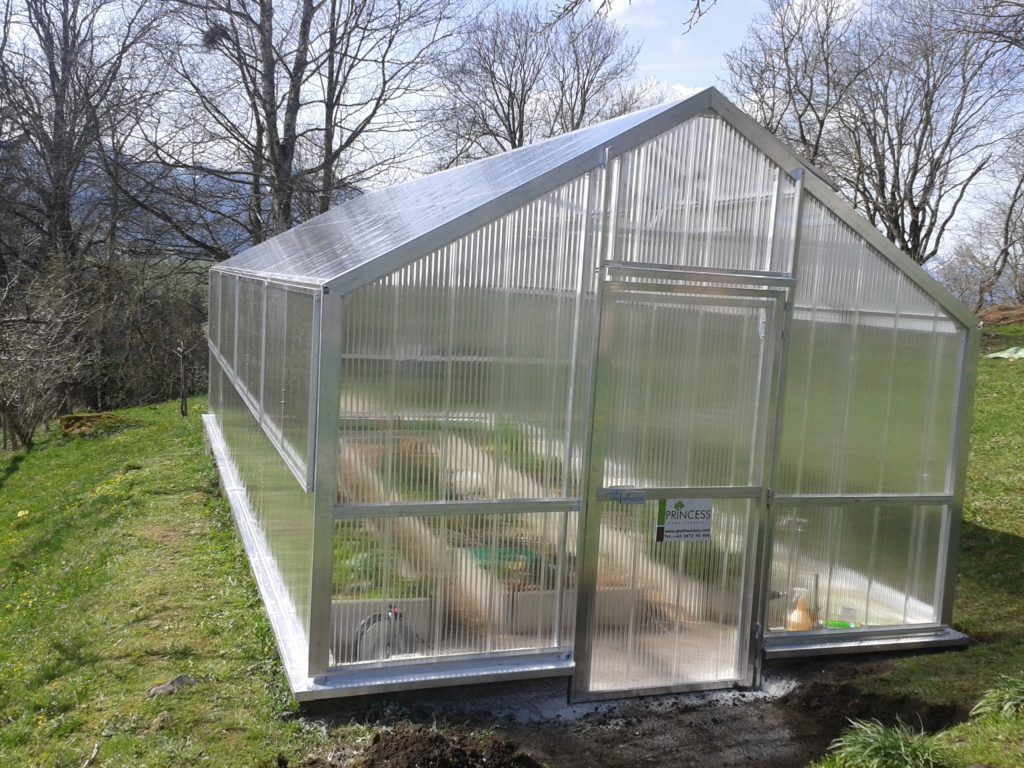 Gewächshaus mit Wänden und Dach aus durchsichtigem Wellkunststoff, an einem grasbewachsenen Hang.