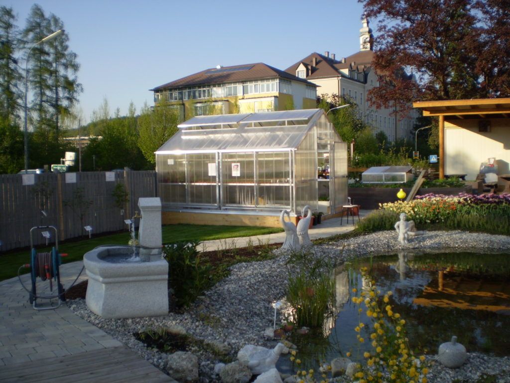 Ein Gewächshaus, ein Springbrunnen und ein Teich in einem Garten mit Gebäuden im Hintergrund.