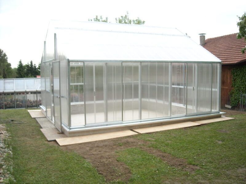 Gewächshaus mit transparenten Wänden und Dach, auf einem Holzsockel in einem grasbewachsenen Hof.