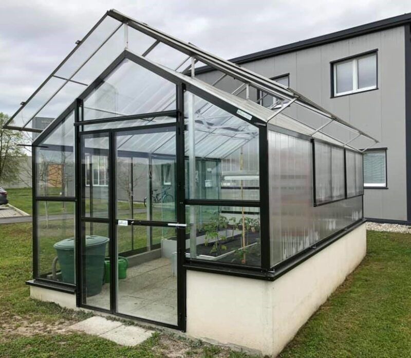 Schwarzes Glasgewächshaus auf Betonsockel, Tür offen, Pflanzen im Inneren, neben einem grauen Gebäude.