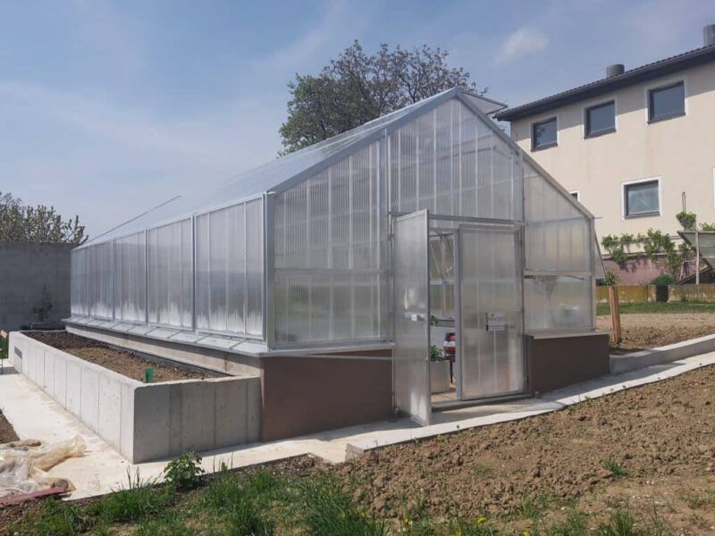 Gewächshaus mit transparenten Wänden und Dach, einer offenen Tür und einem Betonsockel. Braune und betonfarbene Töne.