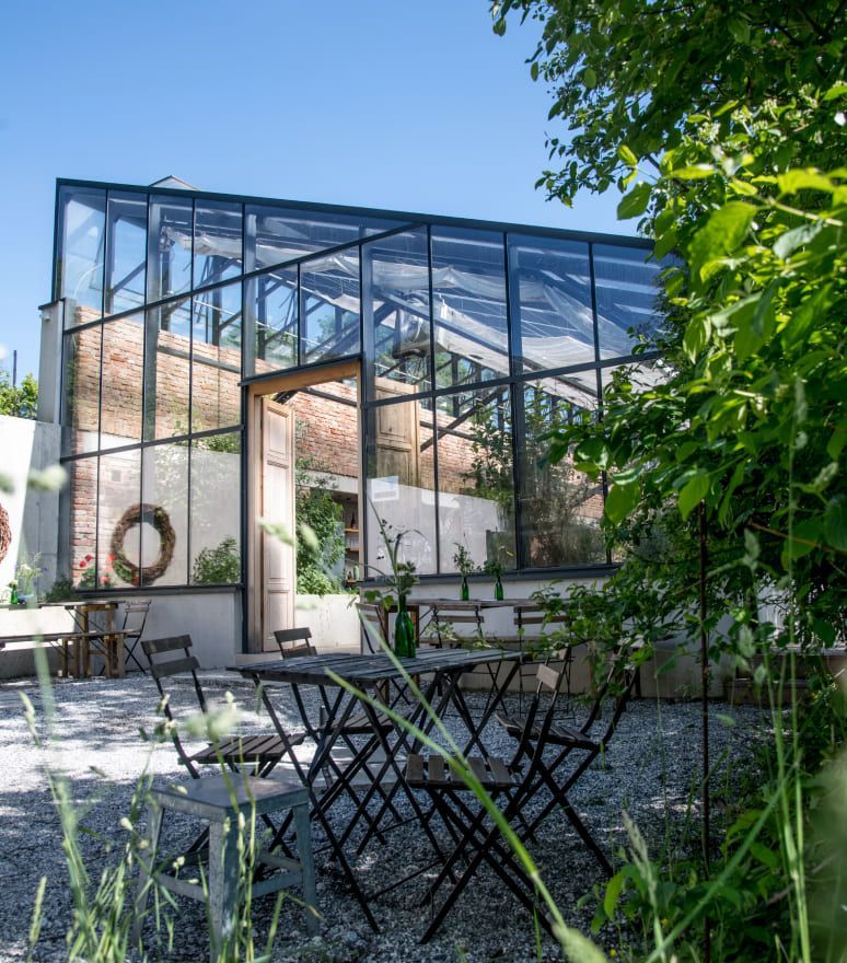 Glasgewächshaus mit Sitzgelegenheiten im Freien auf einer Kiesterrasse. Grüne Pflanzen und Backsteinwände sind im Inneren sichtbar.