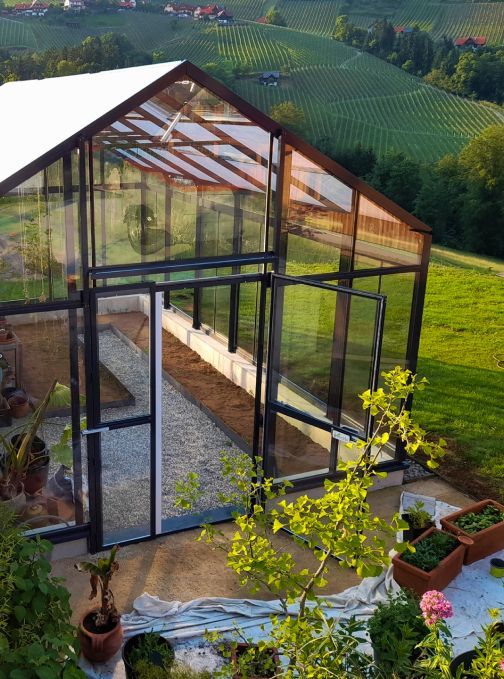 Glasgewächshaus mit offenen Türen, Kiesweg und Pflanzen, eingebettet in einen grünen Hang.