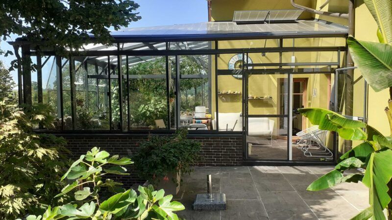 Ein verglaster Wintergarten, der an ein gelbes Haus mit Pflanzen, Ziegelsockel und Terrasse angebaut ist.