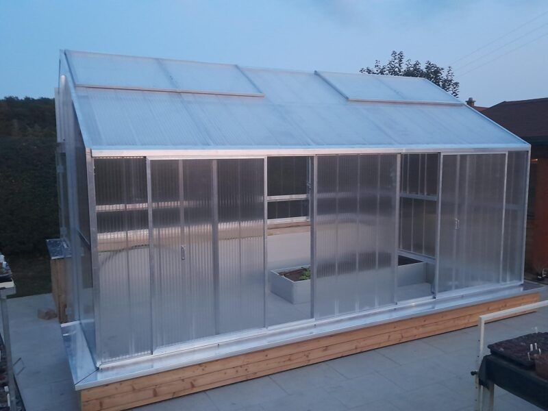 Gewächshaus mit durchsichtigen, gewellten Wänden und Dach, auf einem Holzsockel.