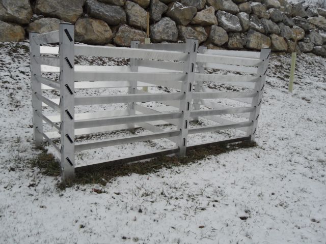 Metallener Baumschutz im schneebedeckten Gras, im Hintergrund eine Steinmauer.