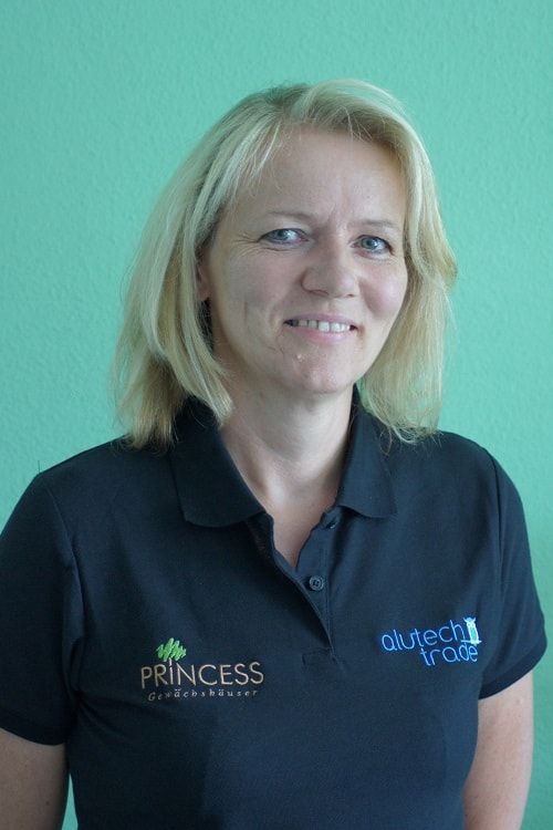 Frau mit blonden Haaren und schwarzem Poloshirt lächelt vor einer grünen Wand.