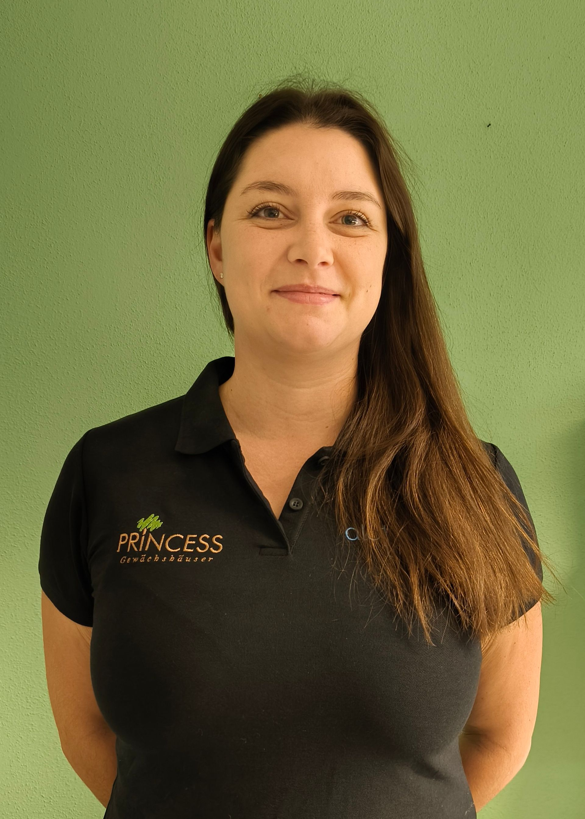 Frau mit Brille und langen Haaren in einem schwarzen Hemd posiert vor einer grünen Wand.