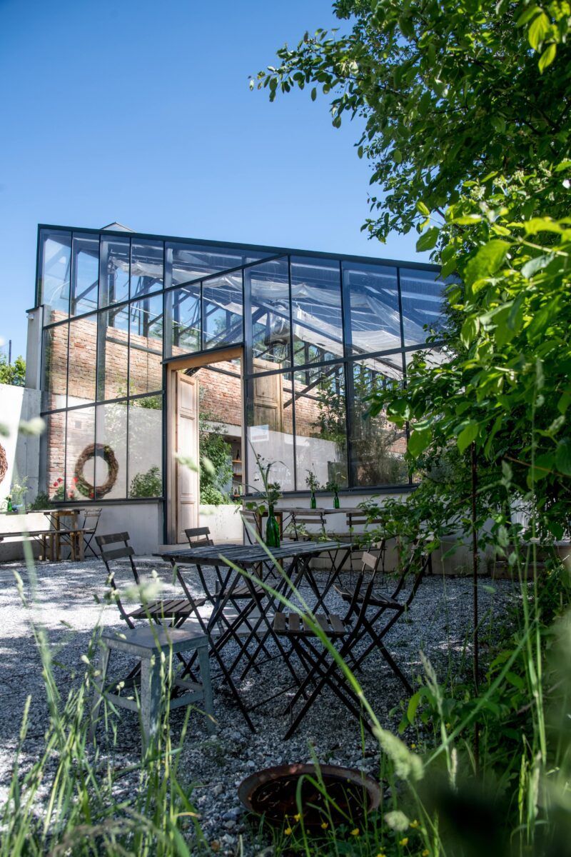 Gewächshaus mit Glaswänden, Backsteinrückwand und Sitzbereich im Freien mit blauem Himmel.
