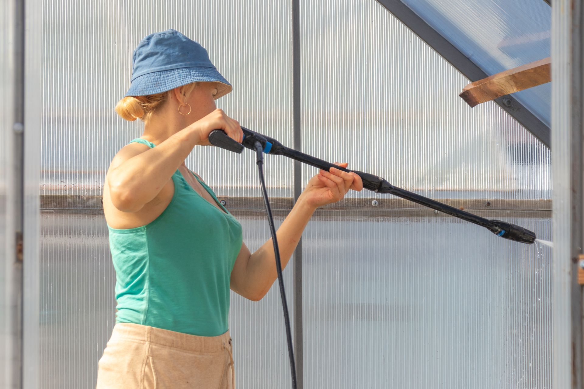 Frau mit Hut benutzt einen Hochdruckreiniger an einem Gewächshausgerüst.
