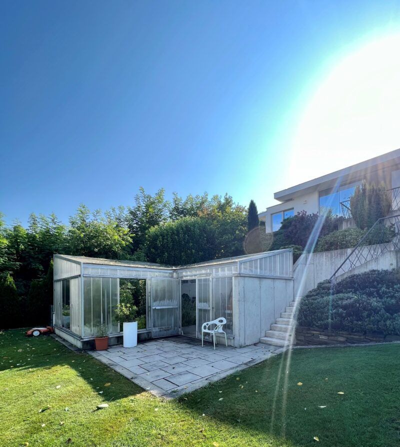 Gewächshaus mit angrenzender Terrasse, gelegen in einem sonnigen Garten mit Rasen und einem Haus auf einem Hügel.
