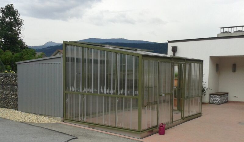 Gewächshaus und Schuppen an einem weißen Gebäude; Berge im Hintergrund.