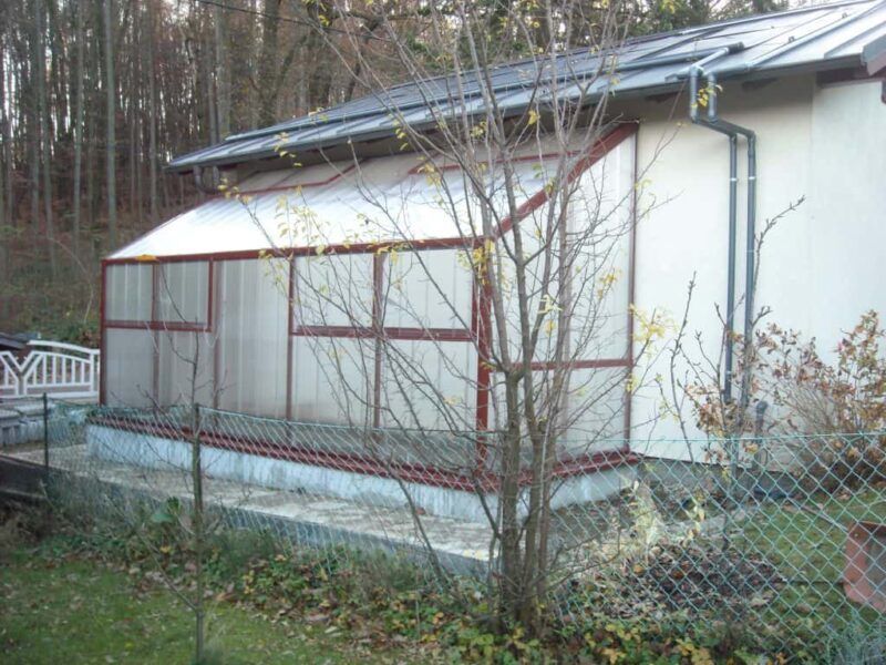 Ein rot umrahmter, mit Kunststoffwänden versehener Wintergarten ist an ein weißes Gebäude angebaut. Er liegt in einem Garten in der Nähe eines Waldes.