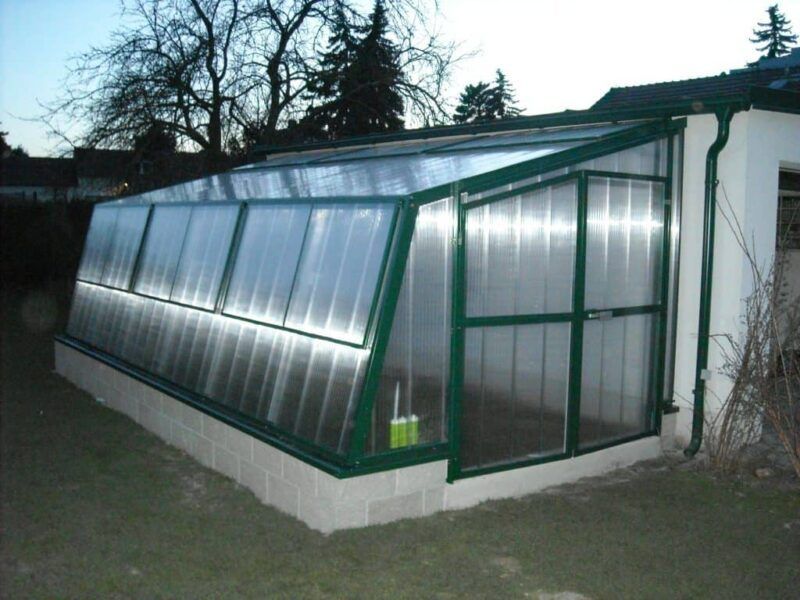 Gewächshaus, das an ein Gebäude angebaut ist und aus einer grünen Metallrahmenkonstruktion und lichtdurchlässigen Paneelen besteht, die auf einem Betonfundament ruhen.