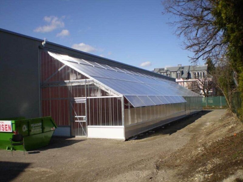 Gewächshaus, das an ein Gebäude angebaut ist und ein transparentes Dach und transparente Wände hat, an einem sonnigen Tag.