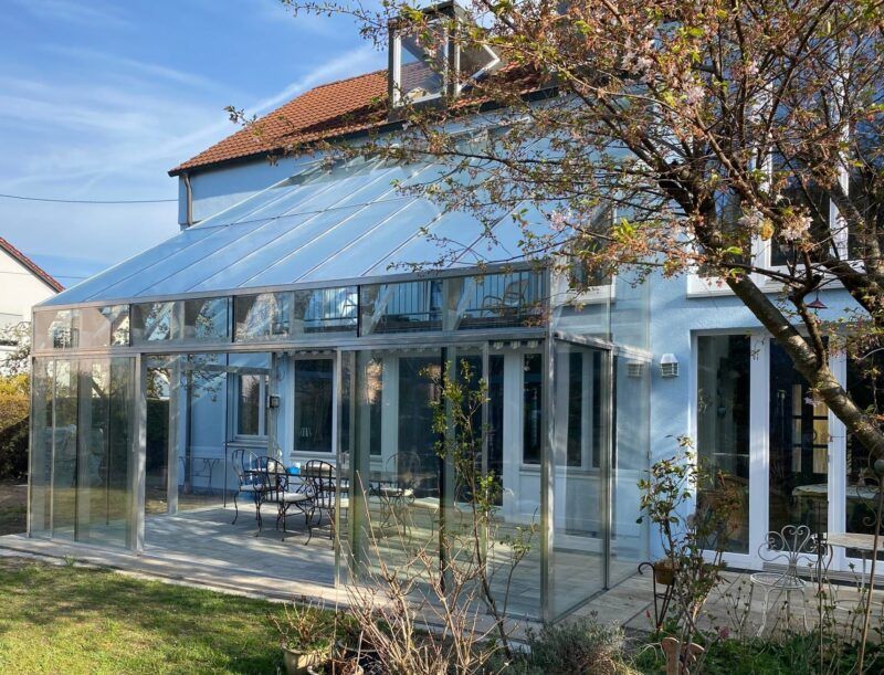 Sonnendurchflutetes, hellblaues Haus mit großem, verglastem Wintergarten und Außenterrasse.
