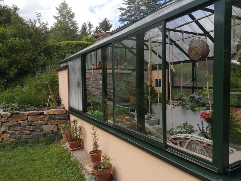 Gewächshaus mit Glaswänden, an einen Hang gebaut. Pflanzen und Blumen befinden sich im Inneren.