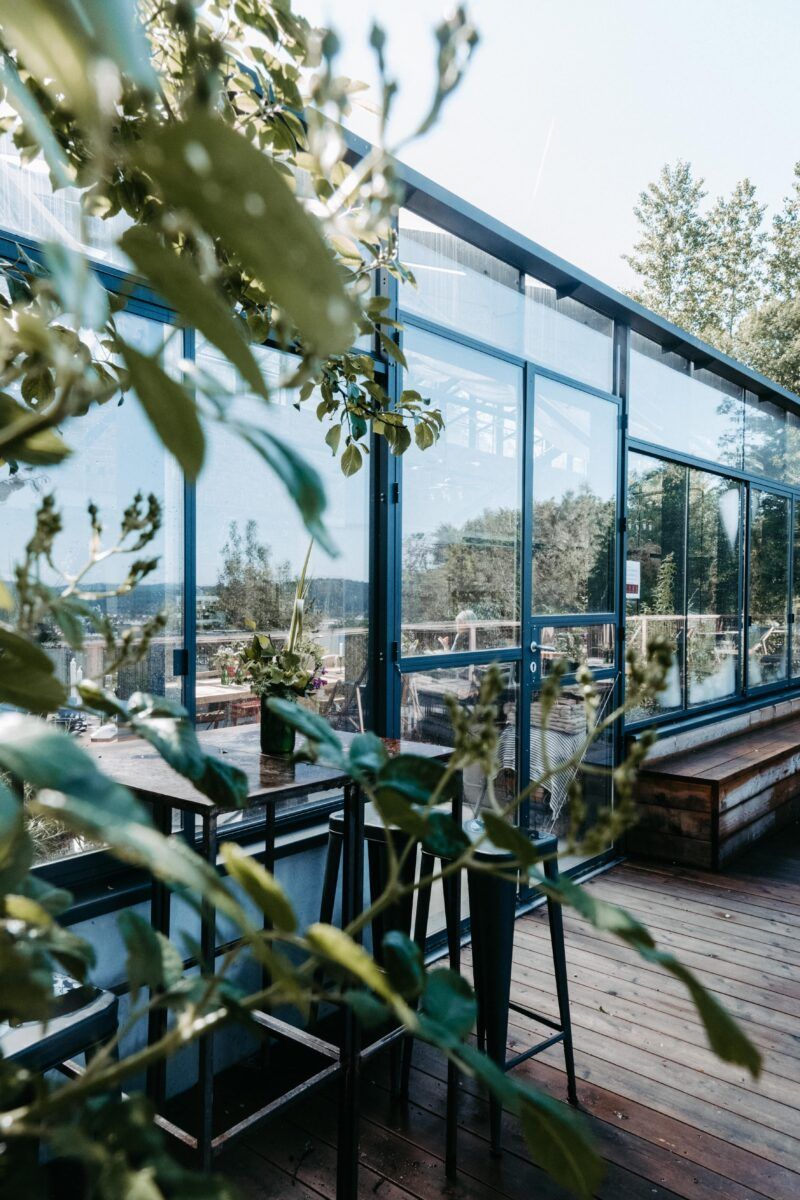 Veranda mit Glaswänden und Blick auf die Bäume, mit Holzdeck, Tisch, Stühlen und Bank.