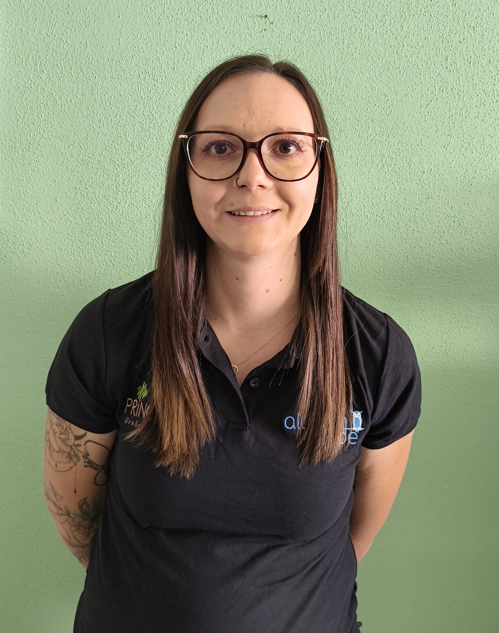 Frau mit Brille und langen Haaren in einem schwarzen Hemd posiert vor einer grünen Wand.