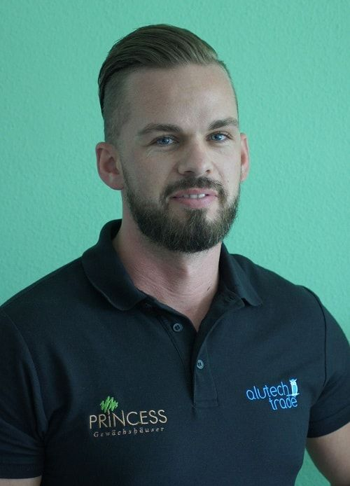 Mann mit Bart und Pompadourfrisur, der ein schwarzes Poloshirt trägt, vor einer türkisfarbenen Wand.