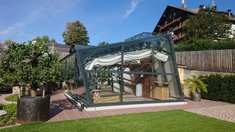 Ein Gewächshaus aus Glas auf einer Rasenfläche mit hölzerner Innenausstattung. Im Hintergrund sind grüne Bäume und ein Gebäude zu sehen.