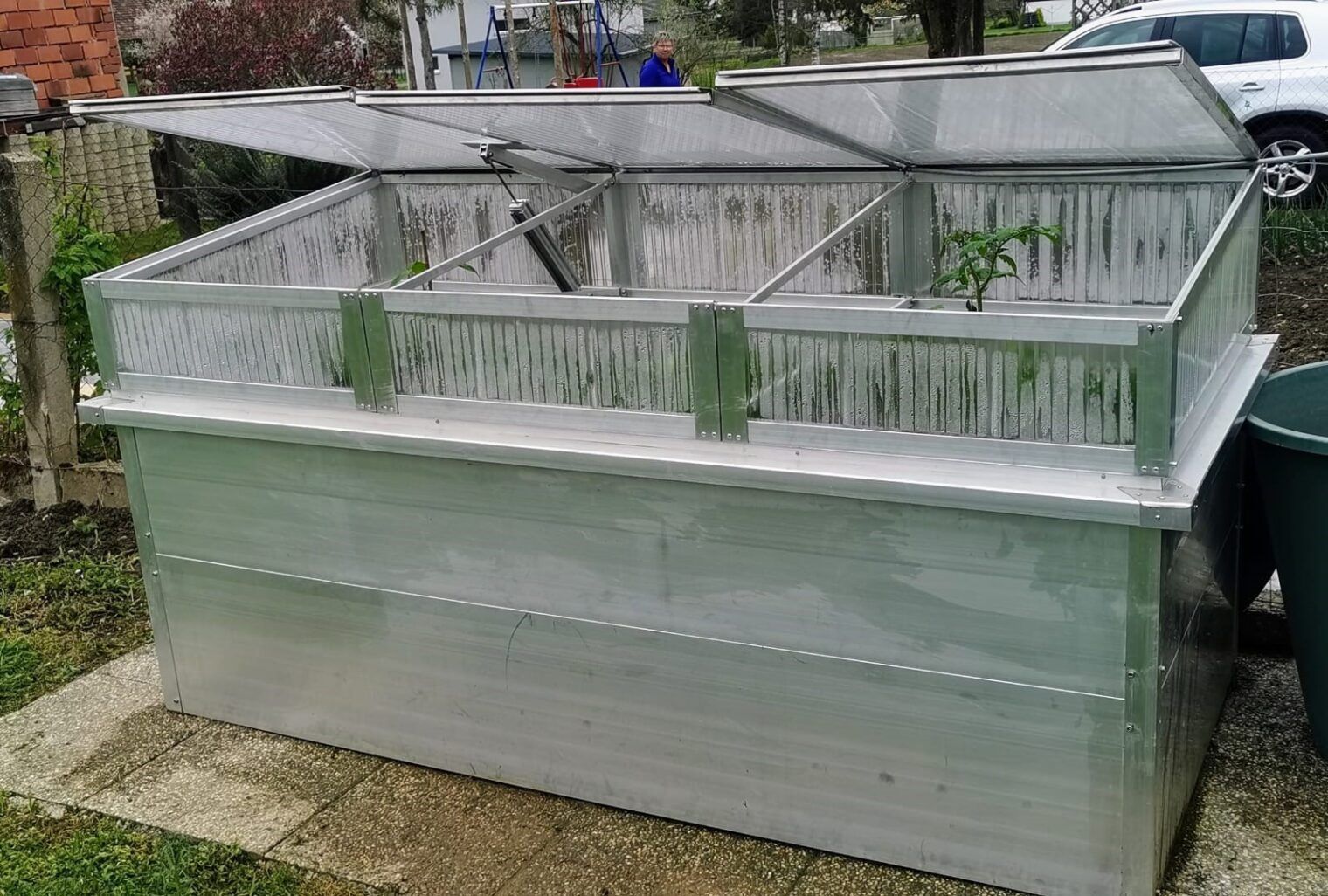 Gewächshaus mit durchsichtigen, aufklappbaren Deckeln zur Aufnahme von Pflanzen, im Freien auf einer Betonplatte.