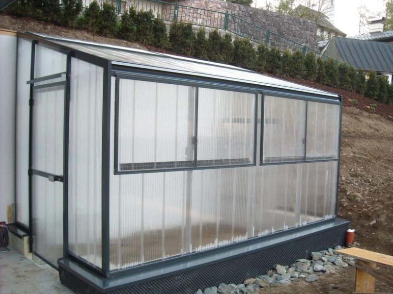Gewächshaus mit durchsichtigen, gewellten Kunststoffwänden und schrägem Dach, in einen Hang gebaut.