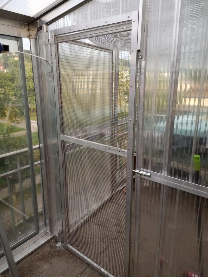 Eine Tür mit Fliegengitter führt in einen Balkonbereich, der aus lichtdurchlässigen Kunststoffplatten und einem Aluminiumrahmen besteht.