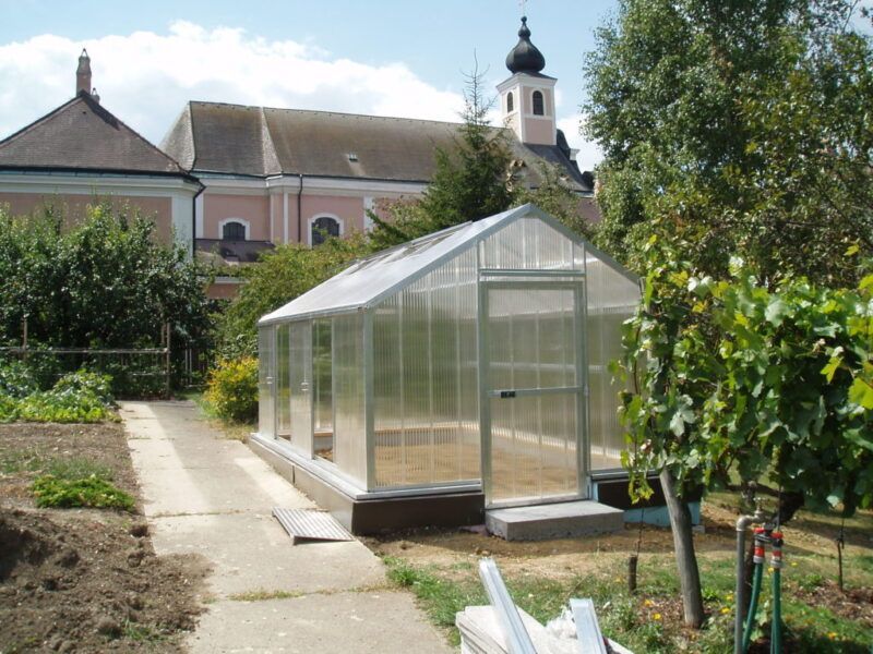 Gewächshaus mit transparenten Paneelen in einem Garten, im Hintergrund Kirche.