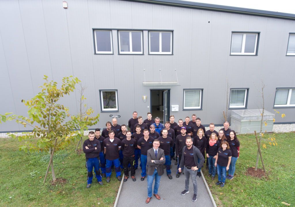 Eine Gruppe von Menschen posiert vor einem grauen Industriegebäude. Einige tragen schwarze Hemden und stehen im Freien.