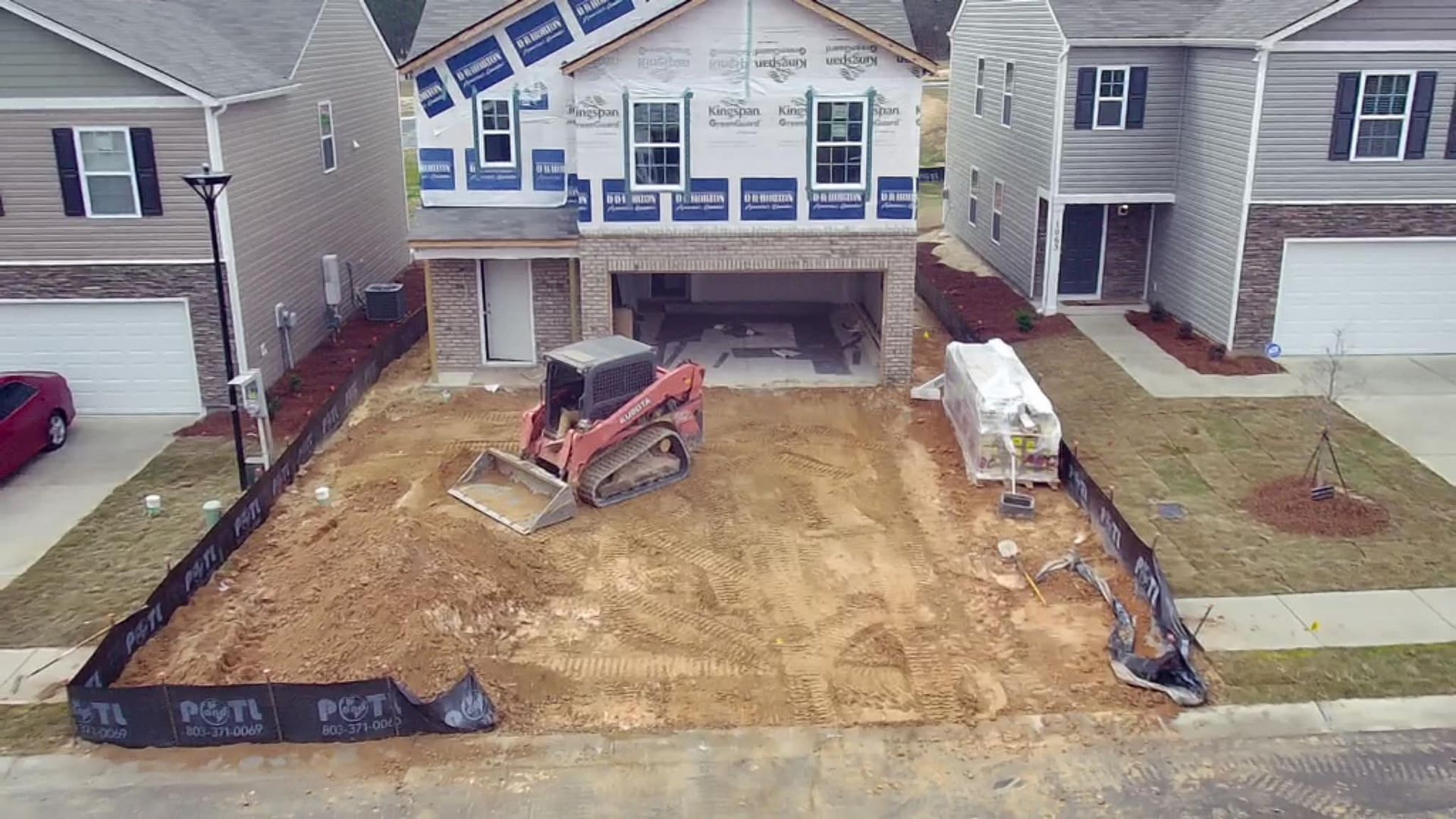 A house under construction with a skid steer on a dirt lot; two other houses on either side.