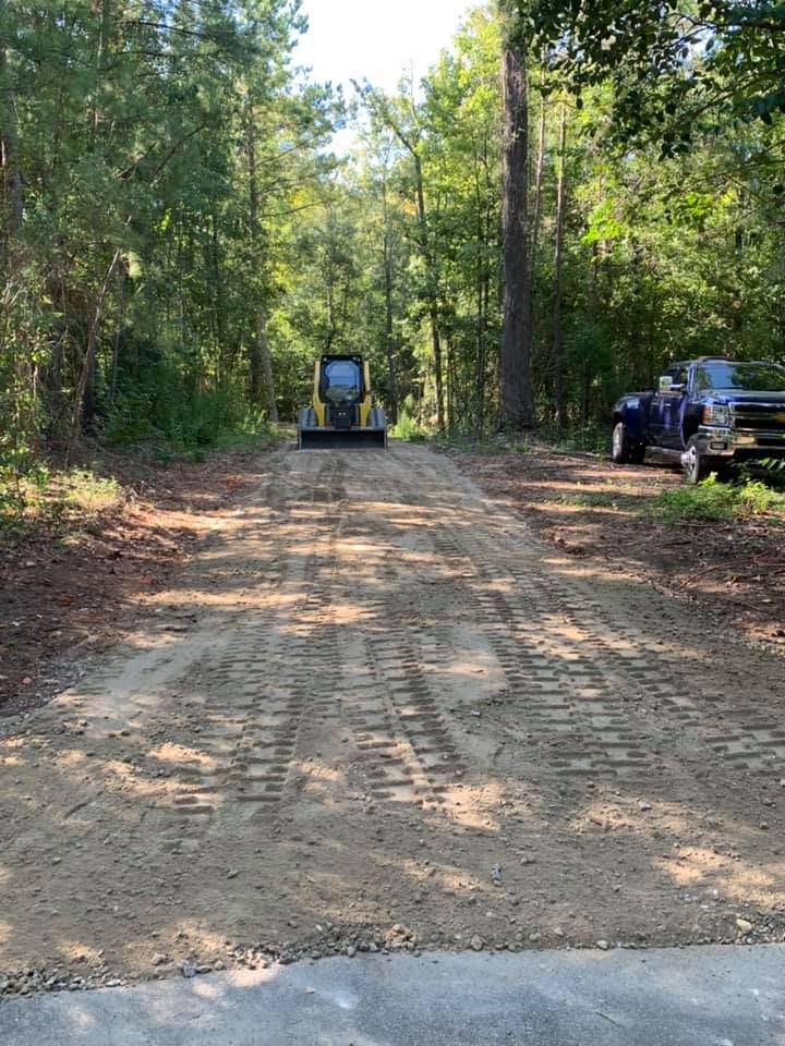Dirt road with a skid steer, trees on either side. A truck is parked on the right.