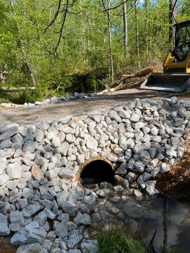 A culvert with stone reinforcement in a wooded area, next to a small excavator.