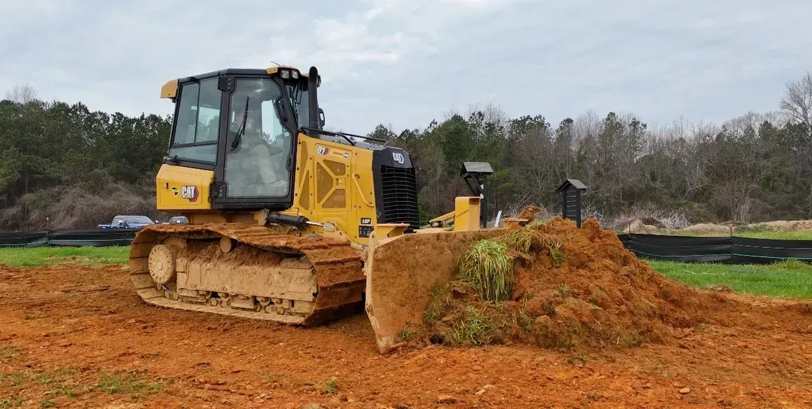 Yellow bulldozer pushing a pile of red dirt on a construction site; trees in the background.