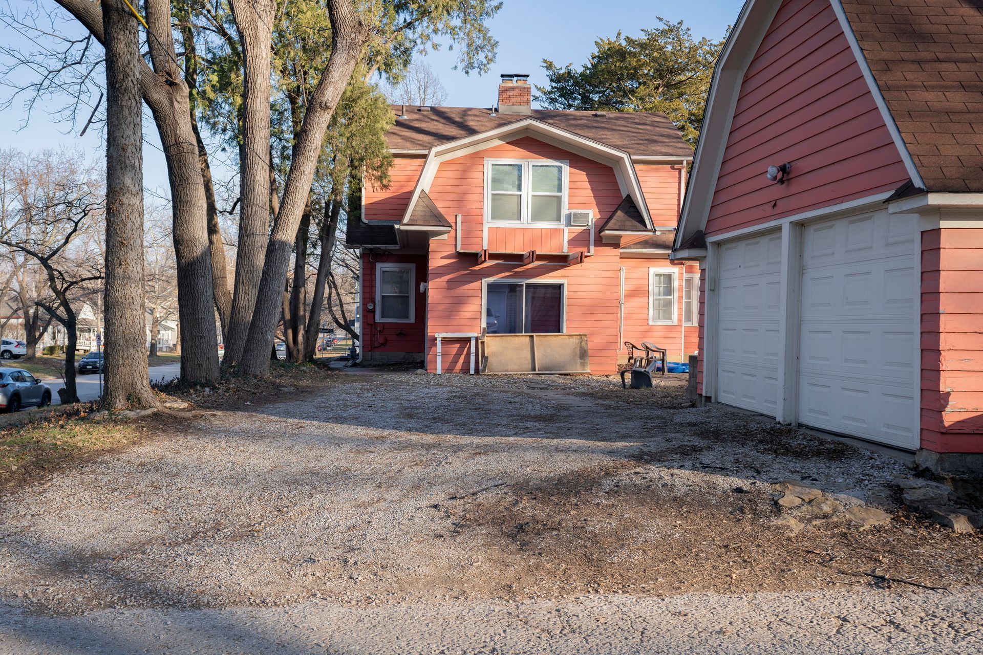 Red house and garage with gravel driveway; trees in front on a sunny day.