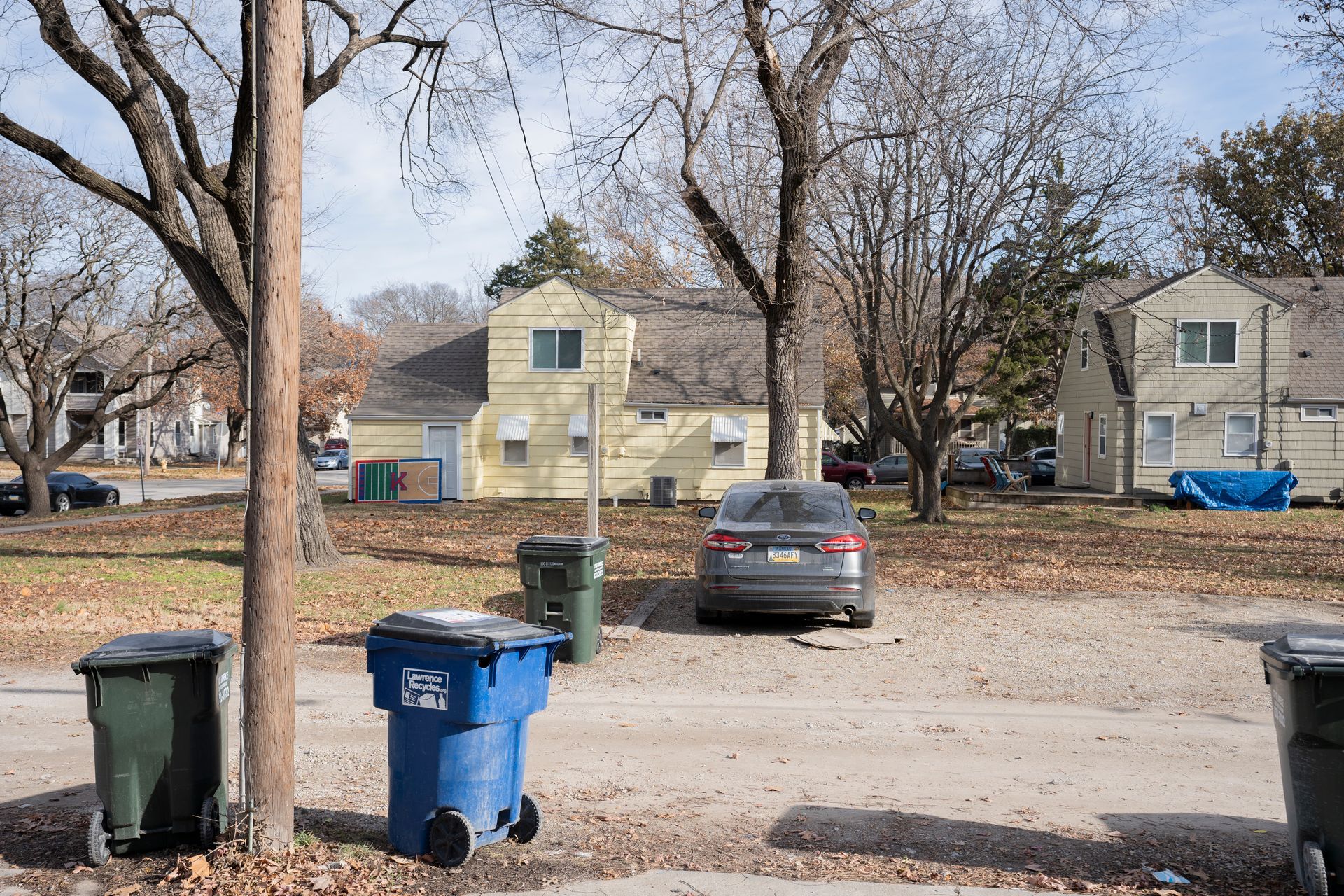 A residential street scene with houses, trees, parked cars, and trash bins on a cloudy day.