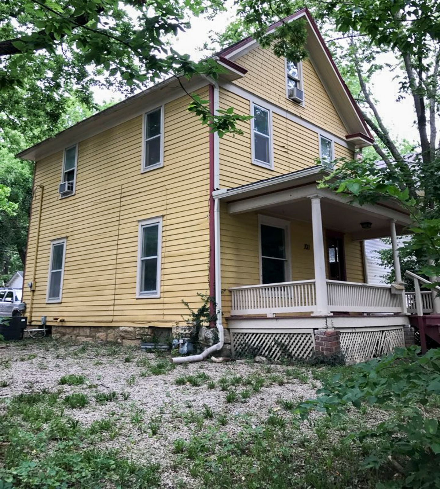 Yellow two-story house with a porch, surrounded by trees and overgrown grass.