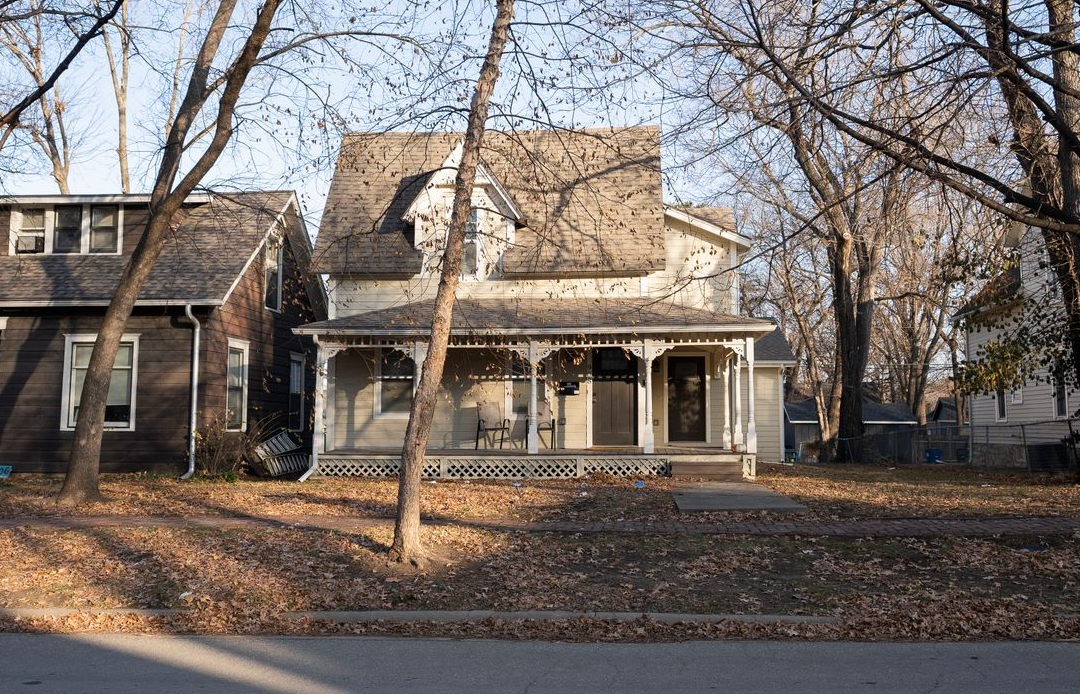 Two-story Victorian house with porch, bare trees, and fallen leaves in yard, on a street.