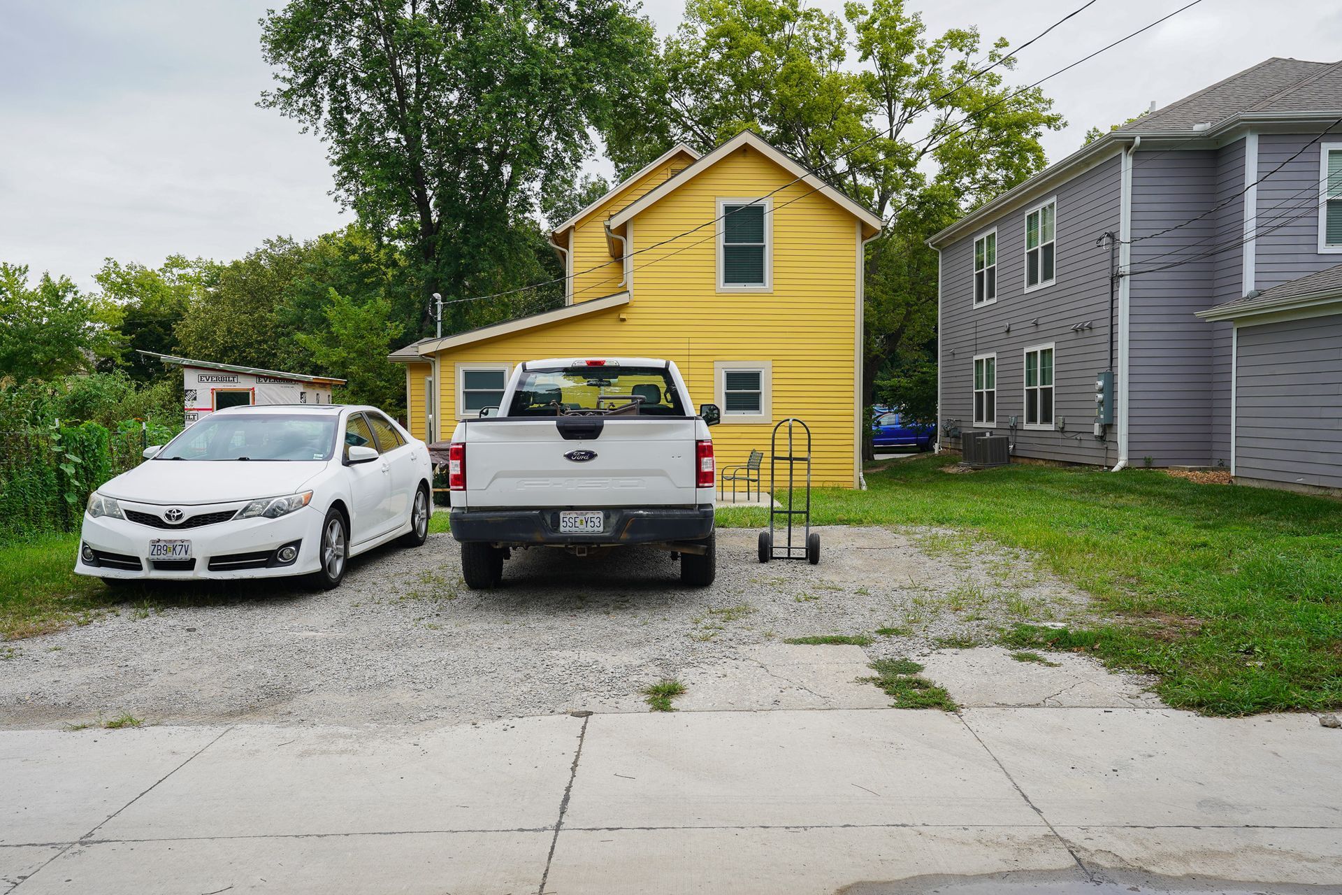 A yellow house with a white truck and car parked in the front yard, next to a gray house.
