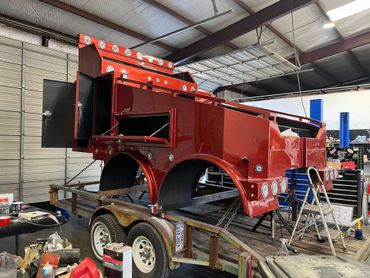 Red truck bed shell on a trailer inside a workshop. Open doors, lights, and tool shelves visible.