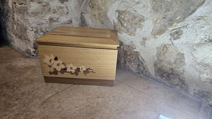 Una scatola rettangolare di legno con intarsi floreali poggia su un pavimento di pietra, addossata a una parete di pietra lavorata.