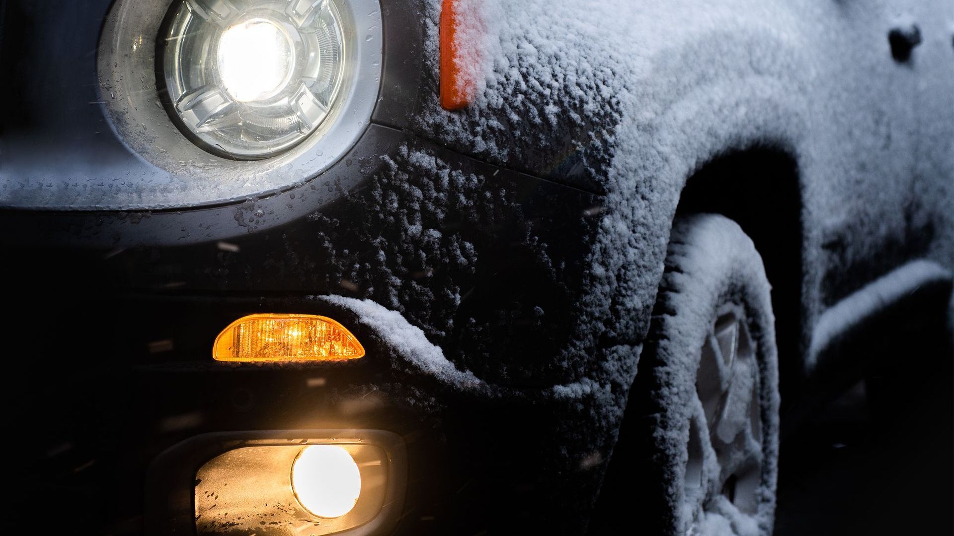 Front of a black car with snow on it; the lights are all on and in working order. 