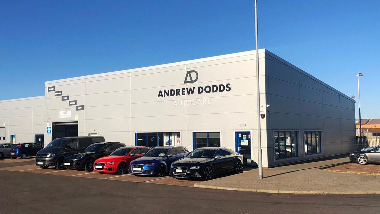 Andrew Dodds Autocare, a car garage in Ayr, in the sunshine with five cars parked out front. The building is large and grey with a sloped roof. 