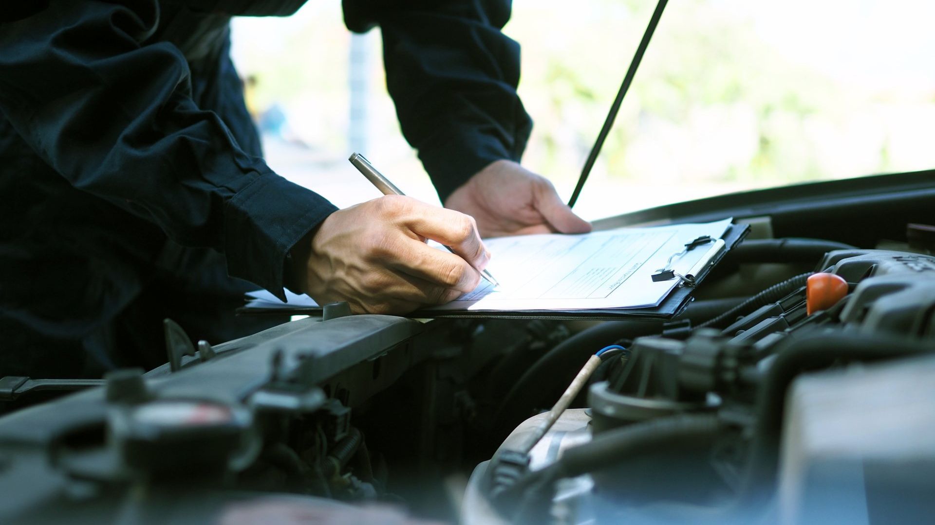 The car hood is open and a mechanic is filling in a form on a clipboard. The mechanic is wearing all black.