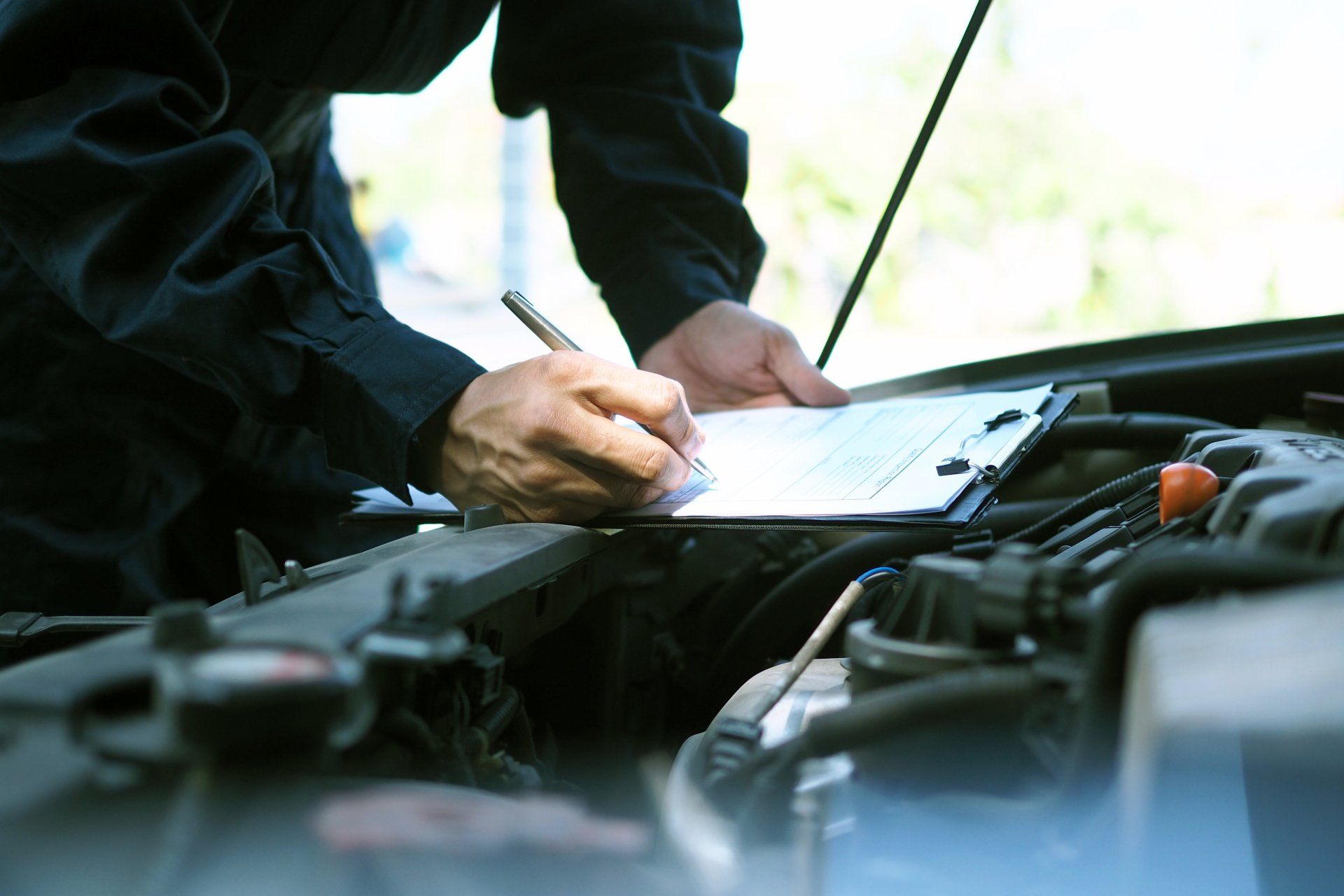 A mechanic is leaning over an open car hood, writing something into his clipboard. The mechanic is wearing all black.