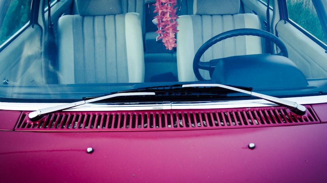 Looking downward on the front hood and windscreen of a pink vintage-style car. We can see the car is empty, and there is a Hawaii lei hanging from the rearview mirror.