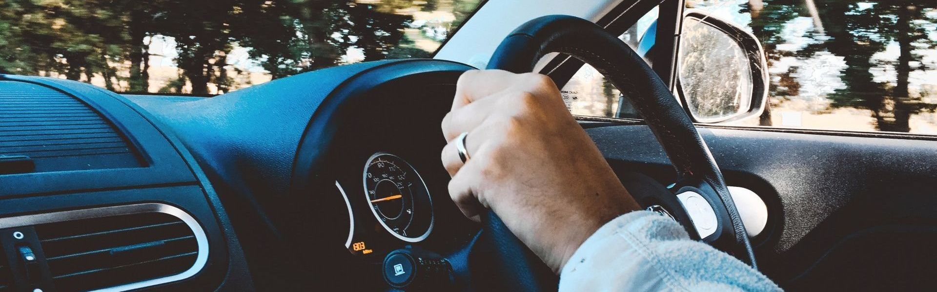 A hand is on a steering wheel, operating a car driving down a wooded road. We can see the dashboard of the car. The hand has a wedding ring on the fourth finger.