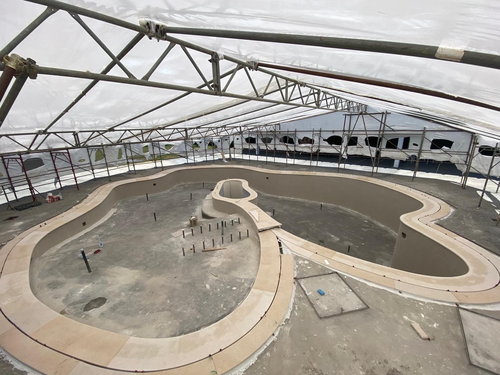 Piscina in costruzione, caratterizzata da una forma libera unica con passerella in legno e impalcature soprastanti.