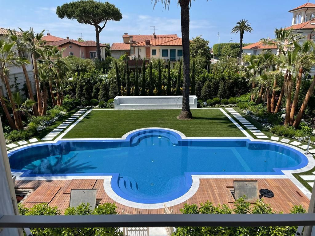 Lussuoso cortile con piscina, prato curato e paesaggio lussureggiante, incorniciato da edifici e alberi.